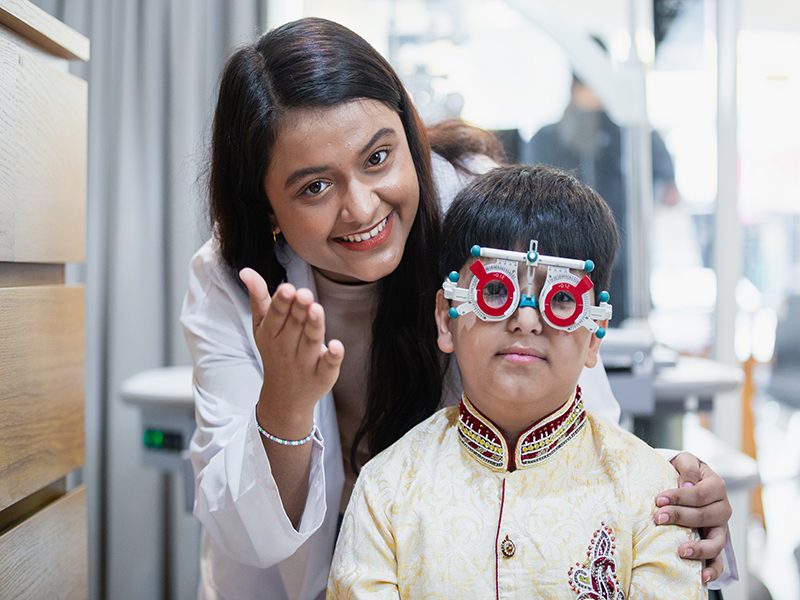 Happy Indian child boy with women eye specialist examining eyesight modern ophthalmology equipment in clinic. Patient kid male checkup iris examines ophthalmological hospital. measure eyeglasses.