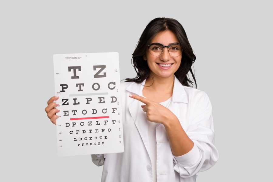 Young indian oculist woman holding an eye chart paper cut out isolated smiling and pointing aside, showing something at blank space.